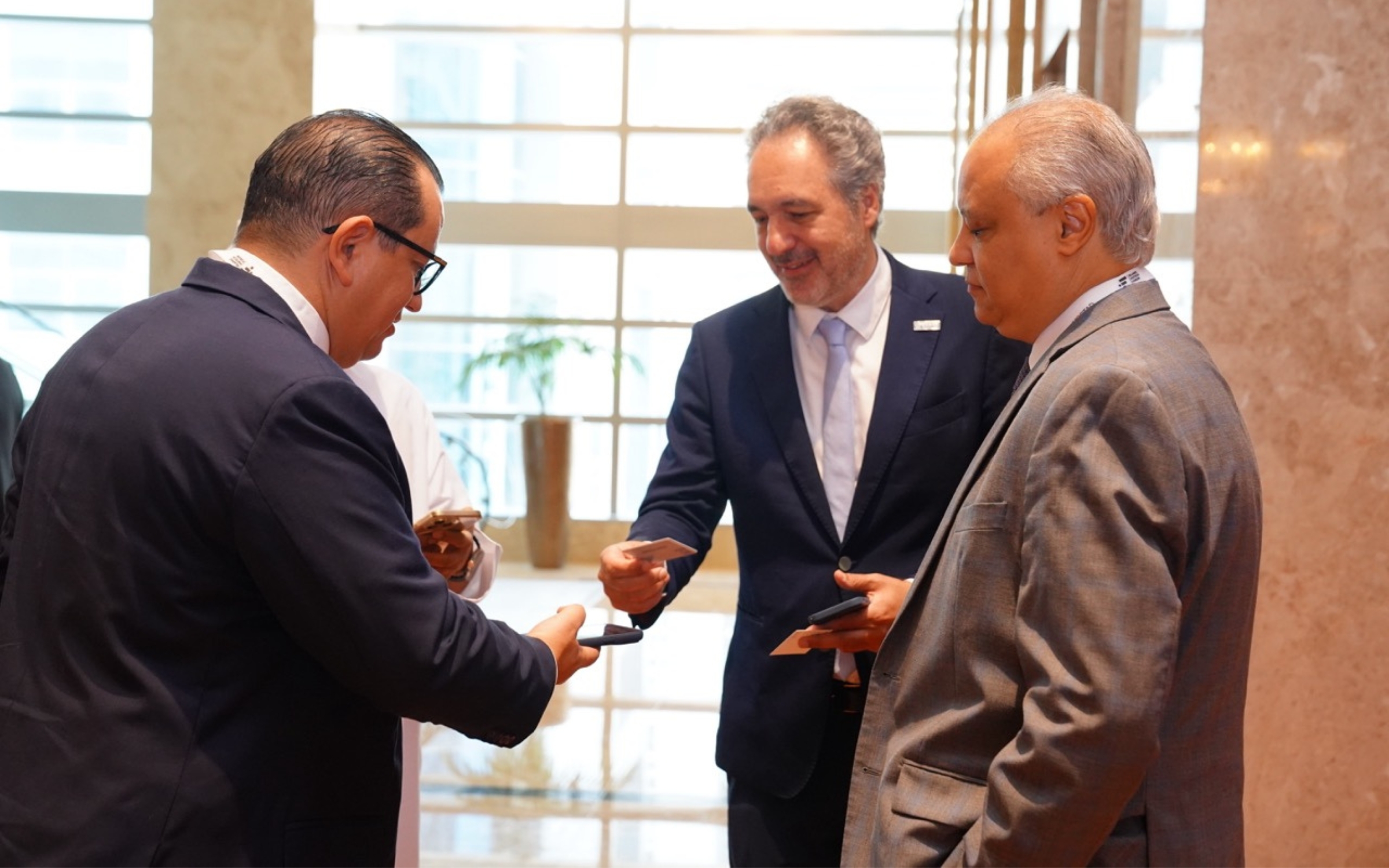 Three senior delegates in conversation, exchanging cards in a conference lobby